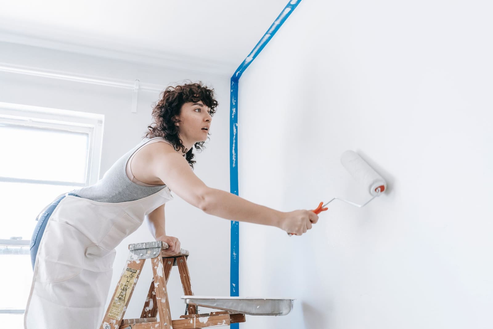 A painter on a stepladder rolling white paint onto a wall.