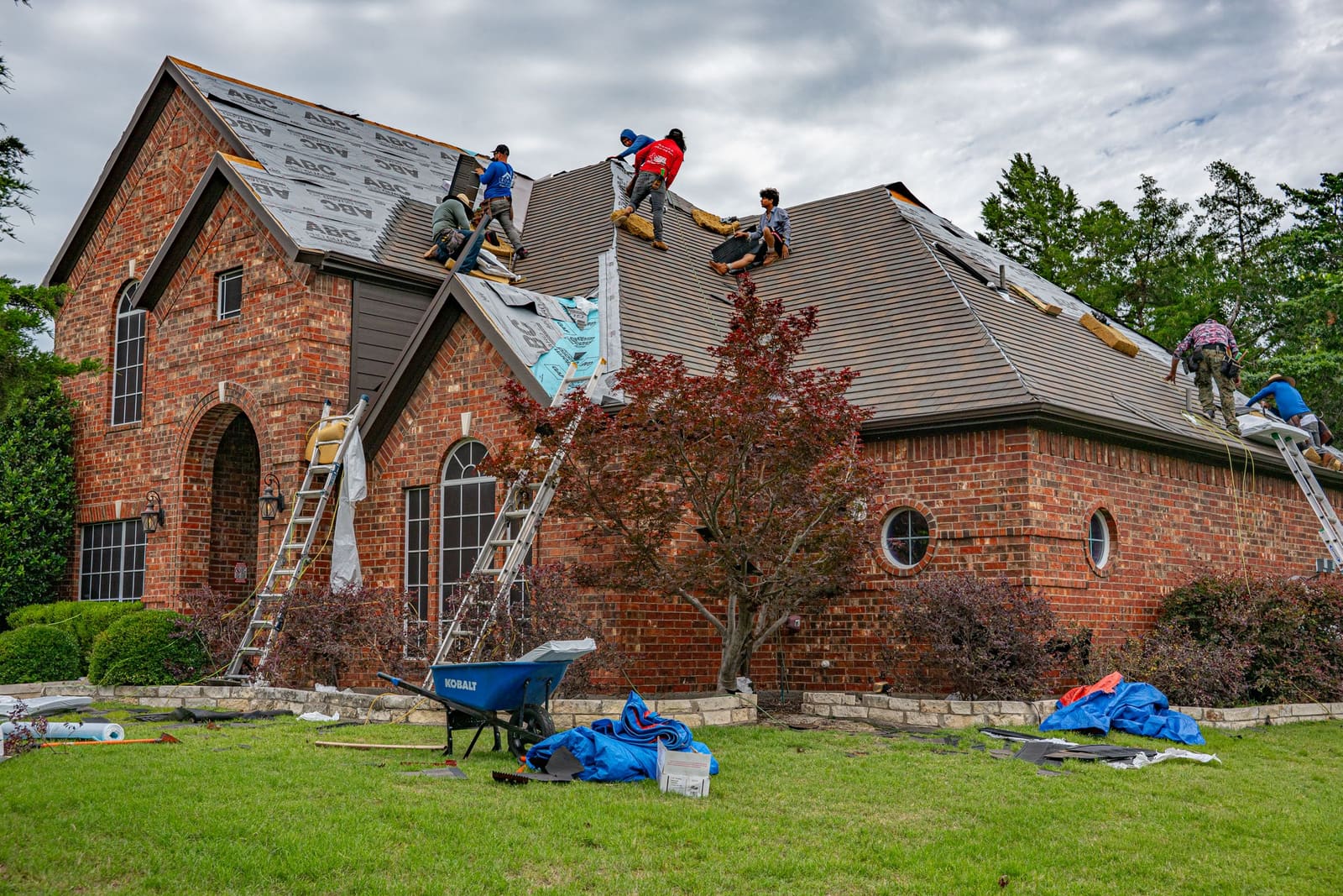 A roofing crew working on the roof of a brick house, ladders up and materials on the lawn.