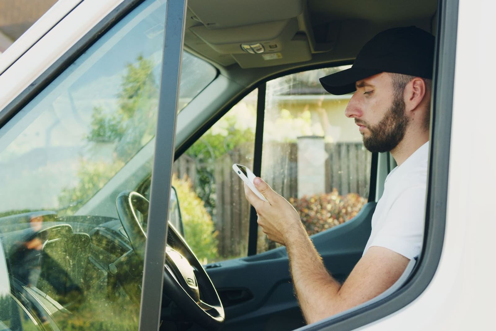 A man in a cap sitting in the driver's seat of a work van, looking at his phone.