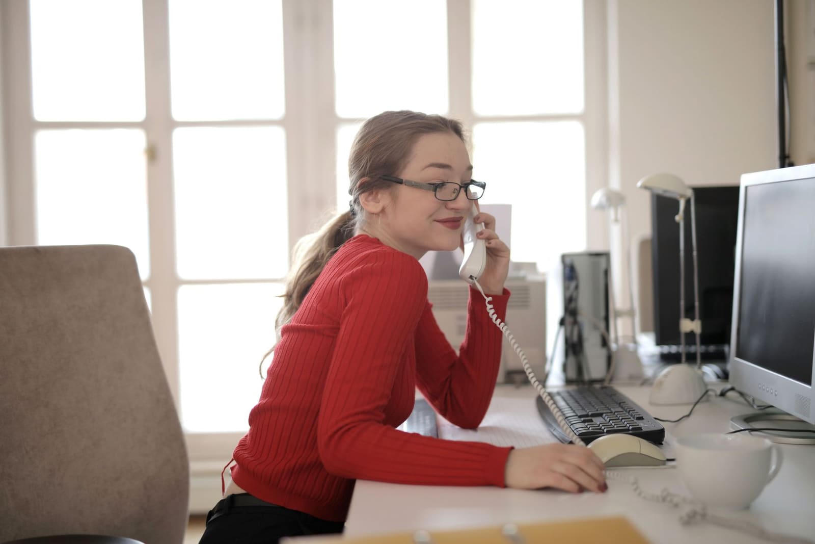 A woman at an office desk, on the desk phone, with a computer in front of her.